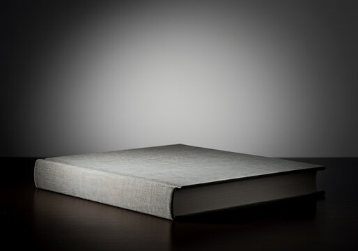 A single, closed, lightcolored book lying flat on a dark wooden surface with dramatic spotlighting against a gradient gray background, evoking a sense of mystery or knowledge