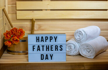  A cozy sauna setting with a sign that reads 'Happy Fathers Day'. There are neatly rolled white towels and a wooden bucket with orange flowers.The concept of spa, relax, rest, health,holiday, gift.