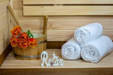 A cozy sauna interior with a wooden bench, a bucket of orange flowers, and neatly rolled white towels. A decorative sign reads Relax. The concept of spa, relax, rest, health, wellness, holiday, gift
