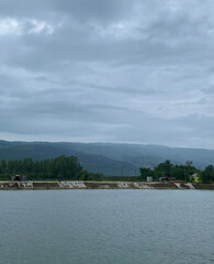 A wide view of a calm body of water against a backdrop of rolling green hills under a cloudy sky