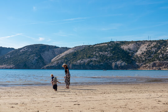 Happy mother playing with her running toddler son on Paradise beach in Lopar, Rab island, Croatia, sharing a joyful bonding moment on wide sandy coast against a backdrop of rugged Mediterranean hills - Powered by Adobe
