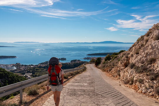 A female hiker carrying a baby in a backpack walks down a steep road towards Rab town. They enjoy the breathtaking view of Adriatic Sea and islands from Kamenjak mountain on a sunny day in Croatia.