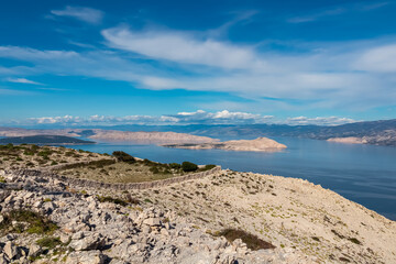 A breathtaking panoramic view from the rocky slopes of Kamenjak mountain looking down at the bare islands and blue Adriatic Sea, framed by the Velebit mountain range under a blue sky in Croatia.