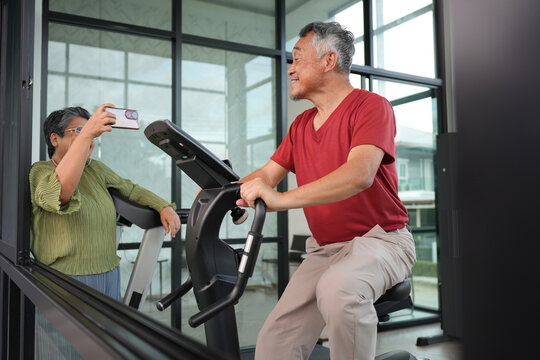 Senior man exercising on stationary bike at home while wife records video, Elderly couple staying active with indoor cycling workout, Active senior lifestyle with older man doing home fitness
