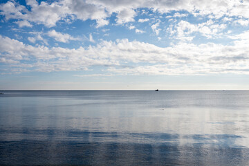 A wide, serene view of the open sea under a vast, bright blue sky filled with scattered white clouds, with a single boat visible in the distance.