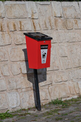 A red dog waste bin mounted on a black pole stands against a beige stone wall in an outdoor area.