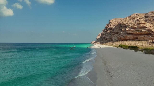 Drone view of stunning coastal landscape with clear turquoise water, soft white sand, and towering rocky cliffs, where small boats create a serene and picturesque seaside atmosphere. Socotra island