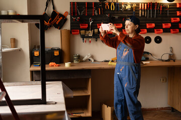 Young professional mechanic or craftsman woman in blue overalls take photo on mobile phone fix work result in repair renovation workshop.
