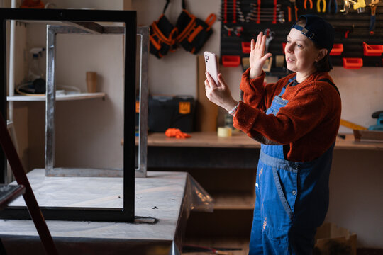 Portrait of smiling woman mechanic or handyman in craft workshop waving on phone during video call.