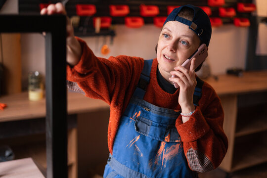 Portrait of female mechanic having phone conversation with customer holding metal construction restoring furniture at workshop.