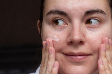 Photo with female portrait сlose up. Mature white woman applies scrub, cream, cosmetic lotion, milk balm to her face with her hands. Concept of beauty, make up, skin care, health, diversity, self care