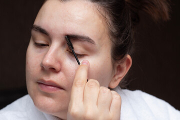 Fototapeta premium Horizontal photo. Female face close up. Mature woman wearing white robe combs her eyebrows with brush. Concept of cosmetic procedure, beauty, make up, skin care, spa, health, routine, rest, relax