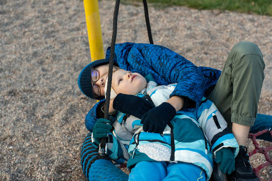 Two brothers lying together on nest swing looking up at sky at playground on autumn day close up portrait. Concept of siblings tender moments, brotherly closeness and peaceful childhood outdoor time.