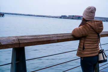 Woman in Brown Puffer Jacket Standing at Waterfront Pier Gazing Over Calm Sea