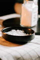 Salt in a black bowl on a table with a glass jar nearby at a cozy setting