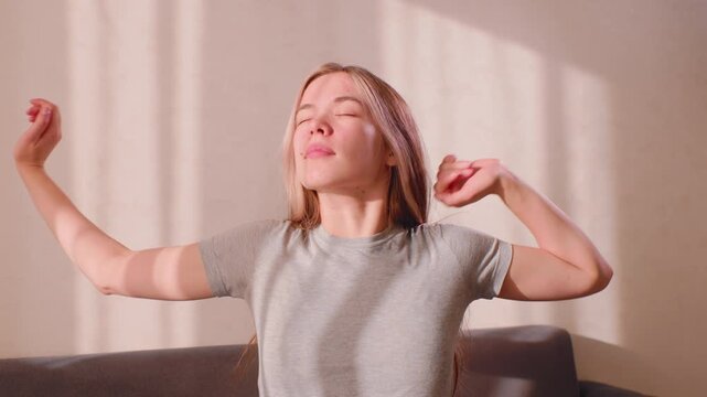 Young woman in grey shirt stretches both arms upward while seated, eyes gently closed, soft sunlight casting warm patterns on wall, expressing calm relief and body awakening