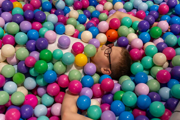 Boy with glasses lying relaxed and buried in colorful plastic balls at ball pit in indoor playground. Concept of childhood sensory play, fun recreation at play center and carefree kids entertainment.