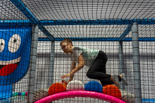  Boy with glasses crawling over large soft balls in netted play structure at indoor playground. Active childhood recreation, physical coordination development and fun at kids entertainment center. - Powered by Adobe