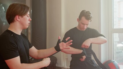 Two athletes wrap hands with black straps in training gym, preparing for boxing or martial arts practice with discipline and concentration, focusing on protection, readiness for workout