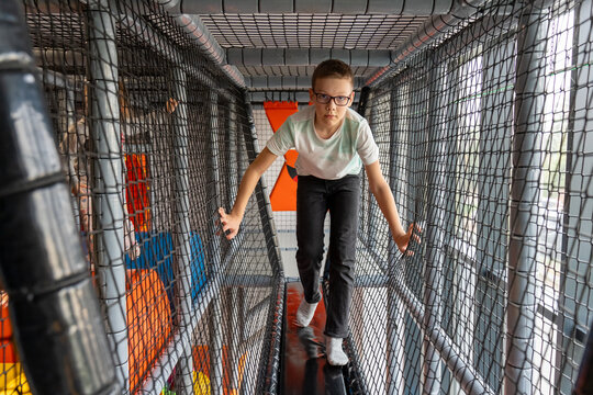 Boy with glasses walking through netted tunnel bridge at indoor playground looking at camera. Concept of active children adventure, obstacle course challenge and fun kids entertainment center.