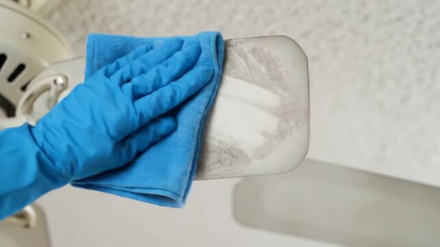 A gloved hand with a blue cloth cleans a dusty ceiling fan blade with a light-colored blade