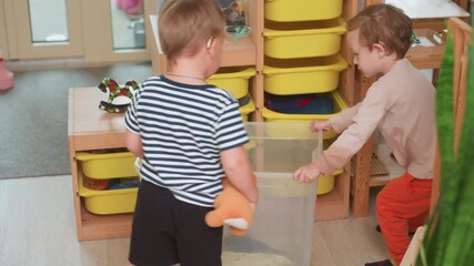 Little boy pushes container with turtle inside while another boy in striped shirt wearing hearing aid holds toy and watches closely during indoor activity surrounded by shelves and colorful drawers