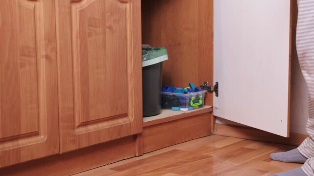 Woman placing transparent box full of plastic bottle caps next to trash bin inside under sink kitchen cabinet, simple home recycling setup, eco friendly household and zero waste lifestyle