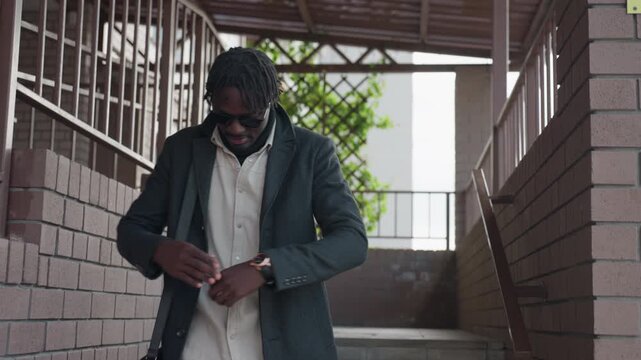 modern man in sunglasses adjusts coat while descending exterior stairs, brick railings and trellis behind, wristwatch glints, confident stride, urban campus entrance with muted colors and modern mood