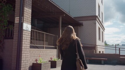female figure exits brick complex into courtyard, long hair flowing, handbag on shoulder, passes vine covered trellis, bright clouds and sunlit walls create urban exterior mood with calm midday vibe
