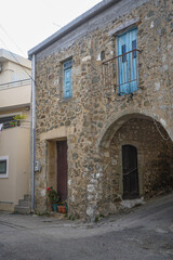 Vertical shot of a historic stone house in Melampes, Crete, featuring an arched entrance, rusted balcony railing, and faded blue shutters, contrasting with a clean, modern building next door.