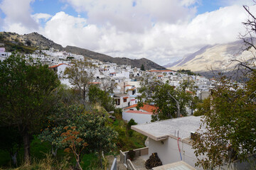 Panoramic landscape of the traditional Melampes village clinging to the mountainside with a dramatic, misty mountain range visible in the distance under a cloud-filled sky.