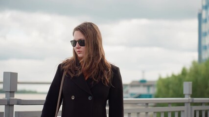 Fair skinned woman in dark coat and sunglasses stands by metal railing, looking toward one direction. Cloudy sky hangs over blue tall building near riverside, breeze moving in soft urban background