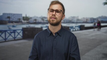 Man blows a kiss with open hand on seaside street promenade near harbor while wearing glasses and beard; playful flirtation.