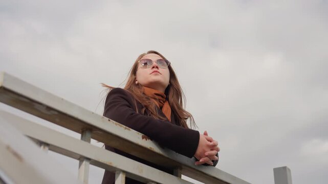 Close view of rusted iron handrail matching tone of cloudy sky with girl in dark suit leaning calmly, reflecting subtle emotion, balance, and contrast between metal texture and soft daylight mood