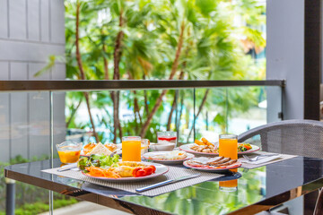 Outdoor breakfast on balcony, tropical garden view. Plates filled with diverse food, glasses of orange juice. Relaxing vacation morning concept, sunny setting.