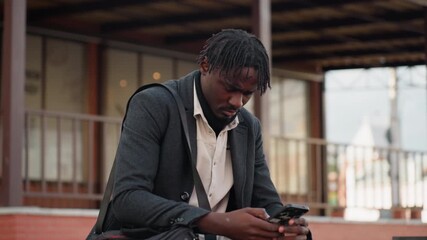 Campus student wearing layered outfit sits outdoors concentrating on phone screen with serious facial expression, showing focus, determination, and digital engagement in calm urban environment