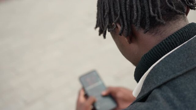 Man holding smartphone displaying unclear images on screen while sitting outdoors in daylight, showing digital technology, scrolling action, and focus on screen content with neutral background tone