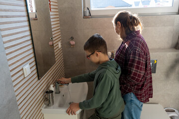 Obraz premium Mother watching son in glasses washing hands at sink in modern bathroom with natural light from window. Concept of parental supervision, teaching children hygiene habits and family morning routine.