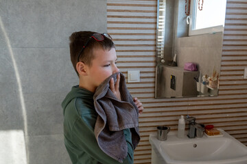 Boy in green hoodie with sunglasses on head wiping face with gray towel after washing in modern bathroom with sink and mirror. Concept of children hygiene, morning routine and healthy habits at home.