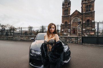 A photograph, a portrait of a beautiful young woman model, a girl in black lingerie, stylish clothes, and a fur coat, standing next to a premium car against the backdrop of an ancient Catholic church.