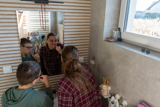 Mother and son brushing teeth together looking in mirror in modern bathroom with natural light from window. Concept of family dental hygiene routine, parenting by example and healthy morning habits.