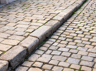 Old stone pavement with a raised curb made of worn cobblestones, showing moss in the joints and signs of ageing along the historic street surface.
