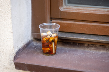 A plastic cup filled with cigarette butts soaked in liquid, left on a window ledge outdoors, showing urban littering and pollution.