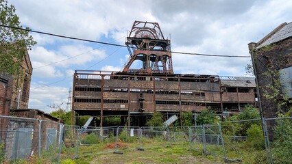 Abandoned coal mining facility, rusting industrial machinery, metal corrosion. Derelict rusty colliery structure historic mine site. Disused ruin fenced off perimeter. Fossil fuels industry in decline