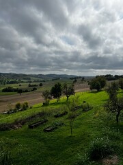 Naklejka premium Dramatic Cloudy Sky over Green Rolling Hills and Terraced Farmland in the Italian or French Countryside with Scattered Trees and Distant Villages