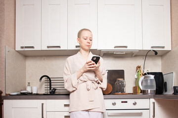 bald woman in a home robe stands near the bright kitchen window looking down at her smartphone,...