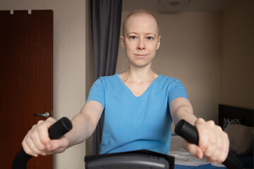 Focused bald woman pedaling on exercise bicycle, cancer treatment side effects and safe home workouts, low impact training for circulation and lungs, rehabilitation
