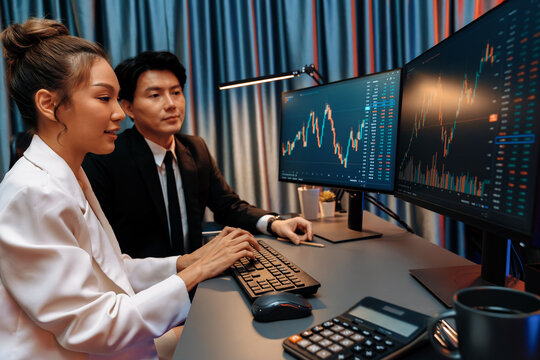 Smiling Asian business traders pointing to dynamic stock exchange data screen profit value online website program at night time at home office. Analyzing currency rate value on data report. Infobahn.