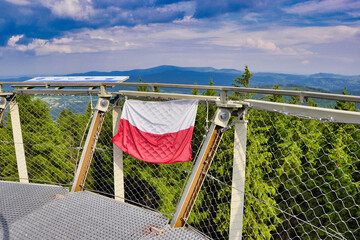 Polska flaga zawieszona na balustradzie punktu widokowego z siatką zabezpieczającą i malowniczą panoramą górską. Widok z wieży w kierunku Gór Sowich. Góry Wałbrzyskie - Borowa. © Pol Badger