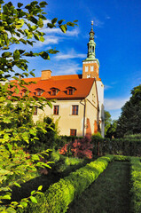 Zabytkowy kościół z czerwonymi dachami i wieżą otoczony zielonymi ogrodami pod błękitnym niebem. Lubiń - Klasztor Opactwa Benedyktynów.